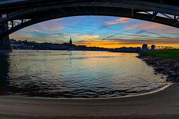 Skyline of the city Nijmegen on the riverbank of the Waal seen from the Waalbrug during sunset. Various ships are passing the river during this beautiful cold winter sunset during the blue hour at the end of the day.