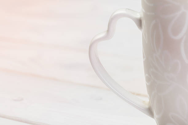 A white mug with a unique heart-shaped handle rests on a light wooden table, softly lit.