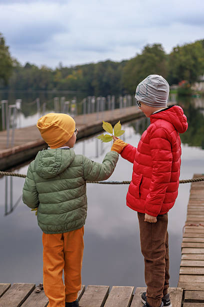 istock Two children in bright jackets exchange autumn leaves while standing on a wooden dock with open copy space. Concept of friendship, childhood interaction, nature and calm reflection. 2250705152