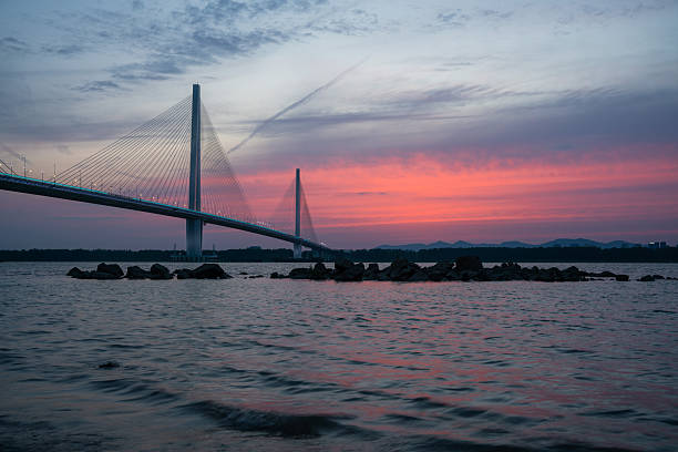 A picturesque sunset illuminates a cable-stayed bridge spanning over water with vibrant colors in the sky.