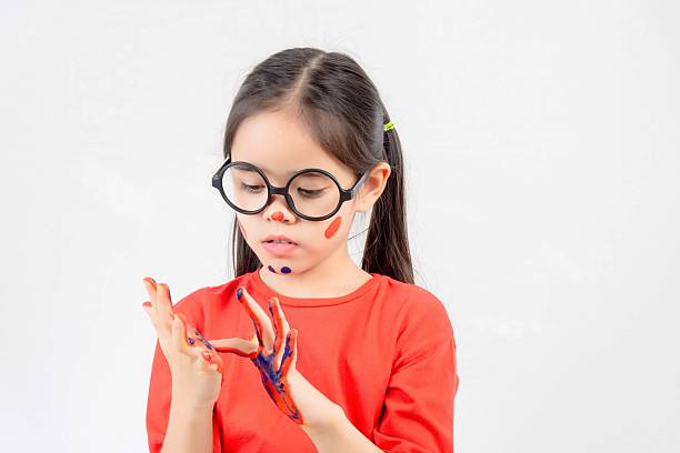 A young girl wearing glasses and a red shirt is painting her hands with blue paint. She looks sad and is holding her hands up to her face