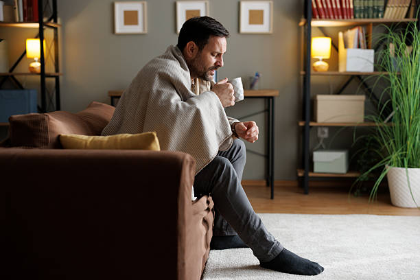 istock Sick Caucasian man wrapped in a blanket drinks warm tea or medicine while resting on a sofa at home 2250715989