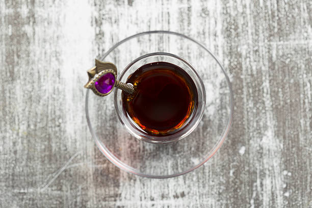 A clear glass cup of dark tea rests on a rustic wooden table, accented by a decorative spoon.