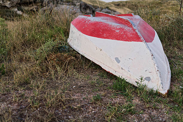 Abandoned Red and White Boat Resting on Overgrown Grass. Abandoned red and white boat lies on overgrown grass in rural setting. Weathered paint signifies years of neglect, surrounded by natural