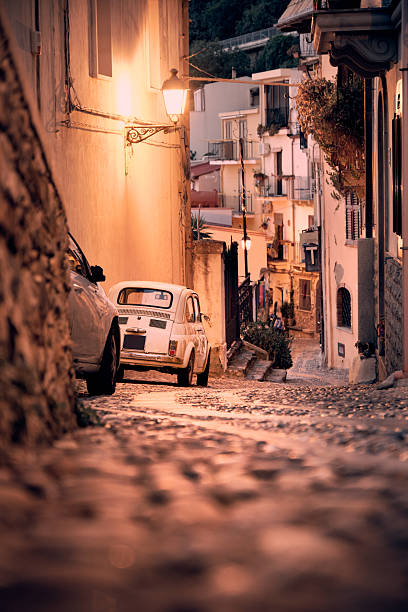 Warm evening scene on a steep cobblestone street in a Mediterranean old town with a small vintage car and traditional houses.