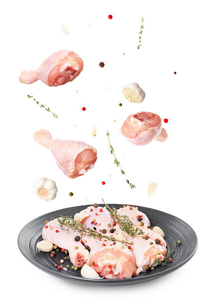 Fresh raw chicken legs with spices and herbs falling onto a plate isolated on a white background.