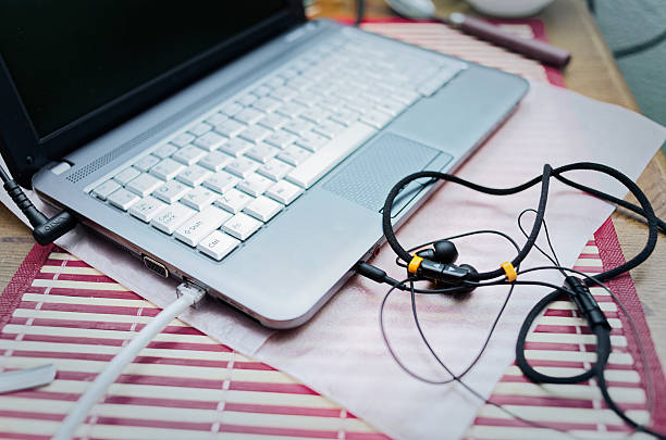 Laptop sits on red and white striped placemat with wired earbuds plugged into its audio jack. Work-from-home setup features blank silver laptop screen on wooden surface.