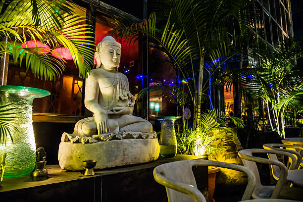A serene outdoor lounge featuring a white Buddha statue surrounded by palm plants and warm lighting, blending spiritual calm with modern decor.