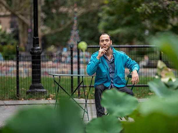 A hispanic man seated at a table in a park, surrounded by greenery and a serene atmosphere, gazes thoughtfully. The background features blurred structures, enhancing the tranquil outdoor setting.