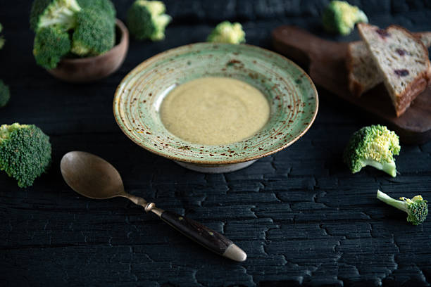 Minimalist food composition with broccoli soup served in a rustic bowl, surrounded by broccoli florets and bread slices on a vintage wooden surface. Cozy homemade meal theme.