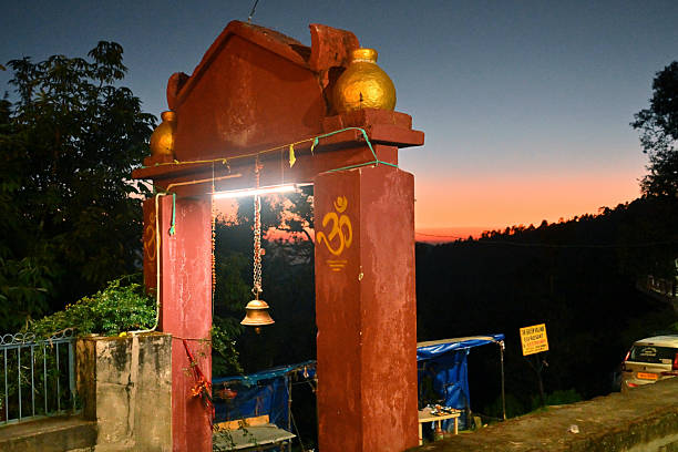 A beautifully lit temple archway stands against a stunning sunset sky at Vriddha Jageshwar, Uttarakhand. The warm twilight hues spread over the surrounding forested hills, creating a serene and spiritual atmosphere typical of the ancient temple complex, where nature and devotion blend harmoniously.