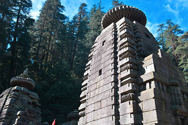 A striking view of an ancient stone temple in the historic Jageshwar Temple Complex, Uttarakhand, India. The intricately carved Nagara-style shikhara rises against a backdrop of towering deodar trees and a clear blue sky. Built between the 7th and 12th centuries, Jageshwar’s temples are revered as one of the oldest clusters of Shiva shrines in the Himalayas. This serene scene captures the spiritual atmosphere and timeless Himalayan architecture of the sacred site.