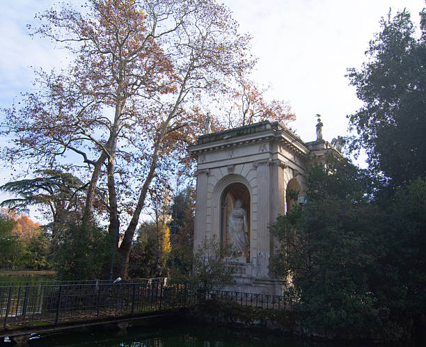 A wide-angle shot capturing the peaceful atmosphere of the Borghese Lake in Rome's Villa Borghese park. The neo classical Temple of Aesculapius stands majestically on a small island, beautifully mirrored in the calm water