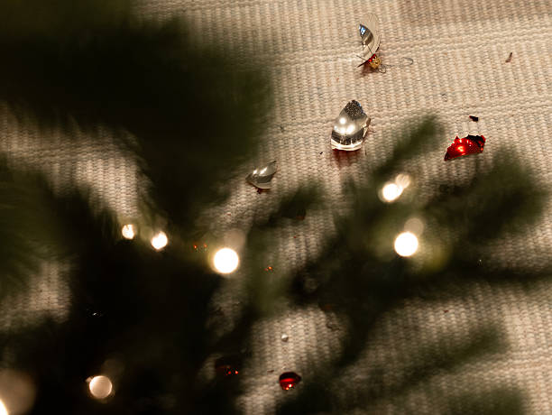 A sad scene of an accident aftermath showing the scattered fragments of a red and silver Christmas ornament in what should be a festive holiday season. With blurred evergreen branches and lights in the foreground, glass shards lay on a beige textured carpet suggesting a fall of the bauble from the tree while decorating it for the winter celebration.