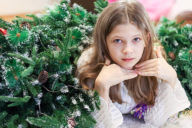 Young girl leans on her hand, surrounded by green foliage, holiday ornaments, and soft lighting in an indoor festive environment.