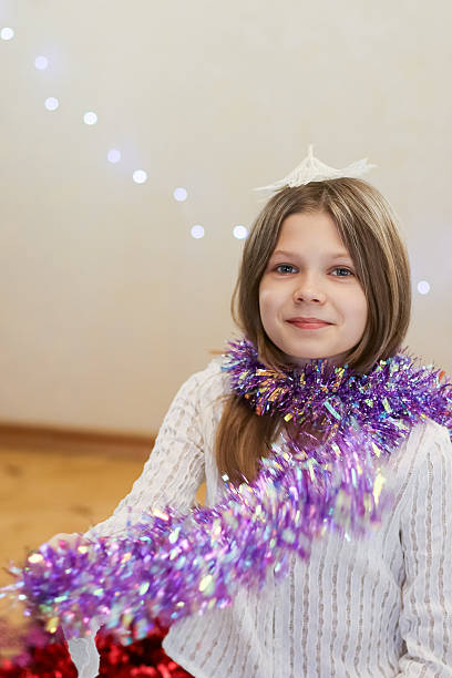 Child wearing white sweater and head accessory smiles while holding out purple shiny garland in room with holiday lights and decorations.