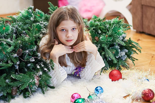 Child lies prone, chin on hands, amid scattered colorful ornaments, tinsel, and artificial evergreens on a soft white surface indoors.