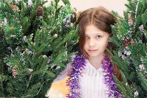 Girl with tinsel necklace peeking out from artificial evergreens dusted with snow and cones