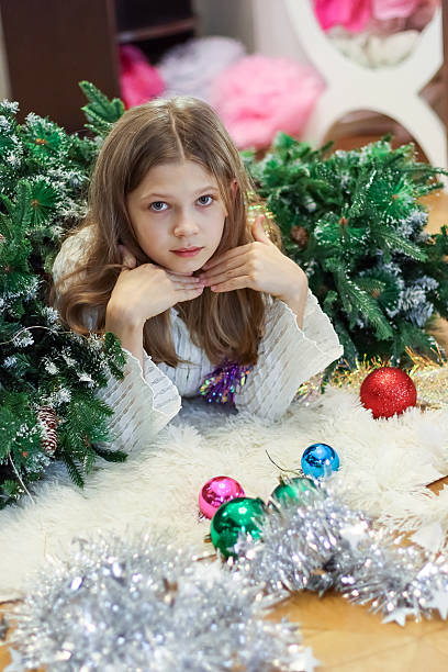 Delight young lady standing near decorated with baubles and garlands Christmas tree on cozy room with star and lights on wall and window.