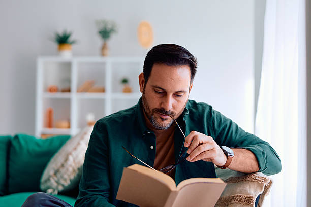 Bearded man holding eyeglasses while reading a paperback on a sofa in a cozy living room, focused and relaxed, enjoying a quiet moment of learning and leisure at home