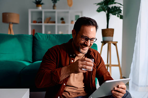 istock Man relaxing home watching tablet, laughing and drinking water 2250777760