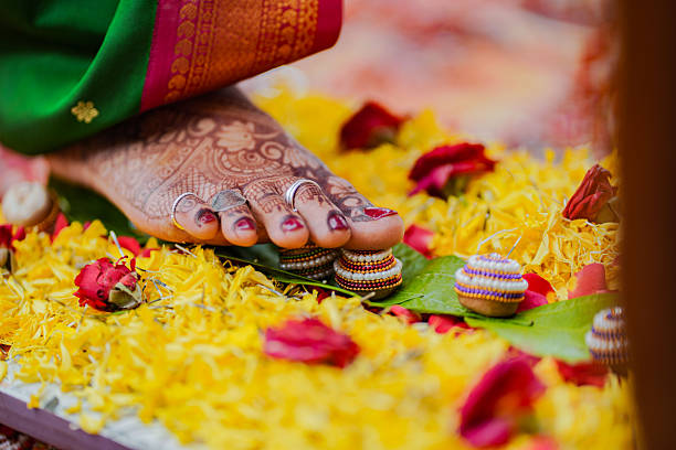 Close-up image showing an Indian bride’s mehendi and toe-ring adorned feet stepping on decorated basari placed over betel leaves and flower petals, capturing an important Hindu wedding ritual.