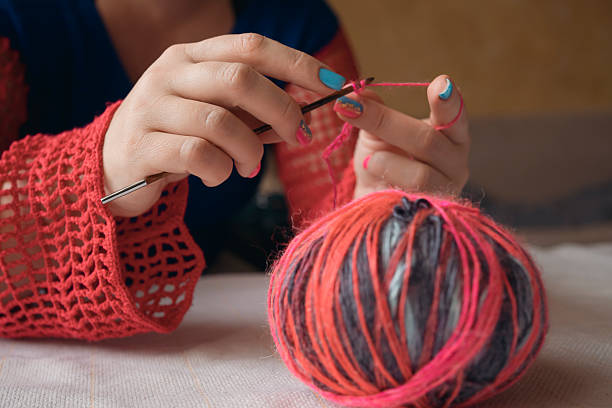 A woman is seated at her desk, crocheting with a ball of purple wool. Her hands are skillfully working with the crochet hook, and the scene captures a moment of creativity in a bright, organized workspace. The desk is neatly arranged with a keyboard, glasses, and a small crochet item, showcasing both productivity and artistic expression.