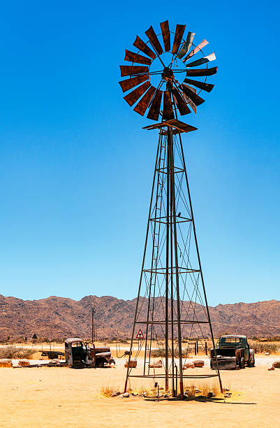 A tall windmill dominates a barren desert scene, with rusted vintage cars scattered at the base and distant mountains under a bright blue sky. Quiet, abandoned industrial landscape conveying heat and solitude.