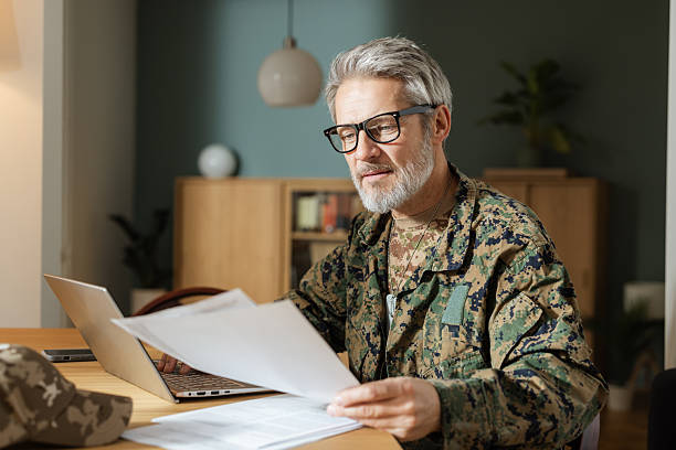 Mature white male military veteran in camouflage uniform working on a laptop and examining papers at home, sitting at a wooden table in a well-lit room with shelves in the background