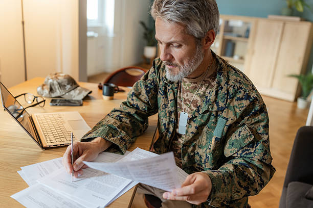 A middle-aged white male military veteran with gray hair and beard sits indoors at a wooden table reviewing documents. Casual home environment with a laptop and shelves in the background