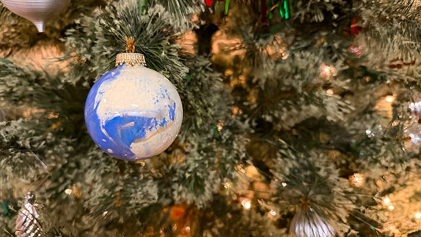 Close-up photograph of a blue and white painted glass Christmas ornament hanging on an evergreen tree with warm glowing lights and soft bokeh creating a festive seasonal ambiance.