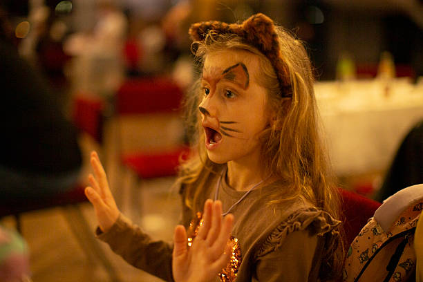 Young girl in cat costume raises hands at indoor party, face painted with whiskers, wearing cat ears and brown dress among blurred crowd and warm