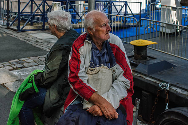 An elderly couple sits back to back on a concrete block near the waterfront in Rotterdam, the Netherlands. The man, dressed in a red and white jacket, gazes thoughtfully into the distance while holding a canvas bag, while the woman, wearing a dark green coat, faces away from him. The scene is set against a backdrop of metal railings, cobblestone pavement, and dockside structures, capturing a candid moment of quiet reflection and everyday life in the port city.