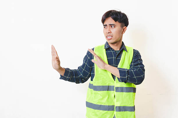 istock Construction worker wearing a safety vest raising both hands in a stop gesture, showing warning and caution on a plain background. 2250817613