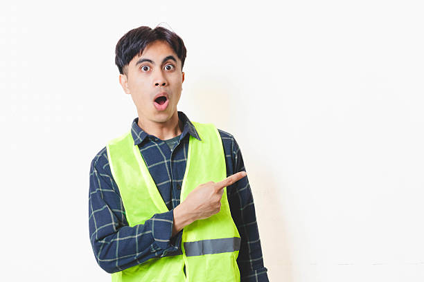 istock Construction worker in a reflective vest pointing to the side with a funny surprised expression against a clean white wall, showing a humorous reaction concept for announcements or attention. 2250817652