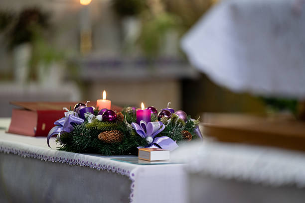 An advent wreath adorned with purple candles, ornaments, pinecones, and ribbons rests on a lace-covered table indoors. Soft candlelight highlights the festive colors while the blurred background adds a calm, reverent atmosphere. The scene conveys warmth, tradition, and the quiet anticipation of the holiday season.
