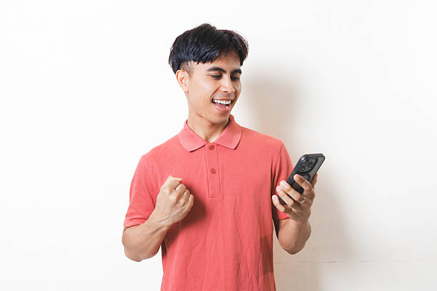 istock Man celebrating success while holding a smartphone, showing excitement and happiness with a cheerful expression on a clean background. 2250823693