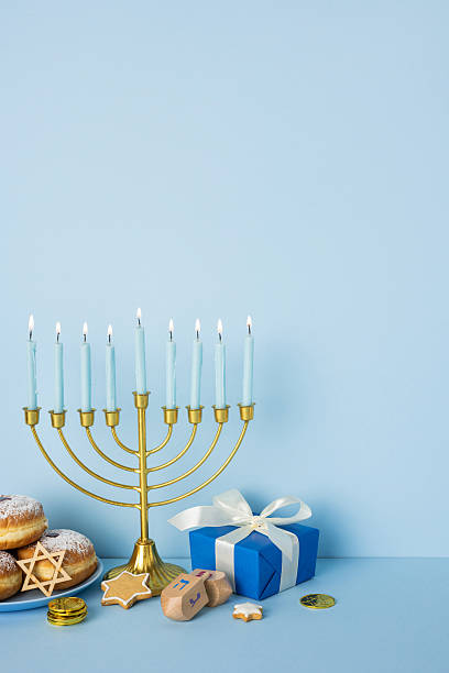 istock Blue vertical background scene features a lit menorah with tall candles, a blue gift box, donuts, latkes, gelt coins, and a dreidel, capturing the warm spirit of Hanukkah celebration and gifts 2250828765