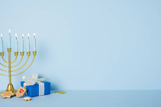 istock Blue background sets a festive Hanukkah tableau: a glowing menorah, lit candles, a blue gift box with ribbon, coins, a star cookie, and other treats, conveying tradition, warmth, and holiday greeting 2250828766