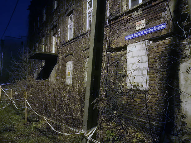 Street-level image captures a contrasting architectural scene with a dilapidated, brick building at the center, flanked by two well-maintained, light-colored apartment buildings. The central building shows signs of age and neglect, with boarded-up windows and graffiti on its lower level, creating a sense of urban decay. In the foreground, a multi-lane road with traffic signs and yellow bollards suggests an active city street. The sky is overcast, contributing to a somewhat somber atmosphere. The juxtaposition of the old, decaying structure with the newer, restored buildings highlights the layered history and ongoing transformation of urban environments.