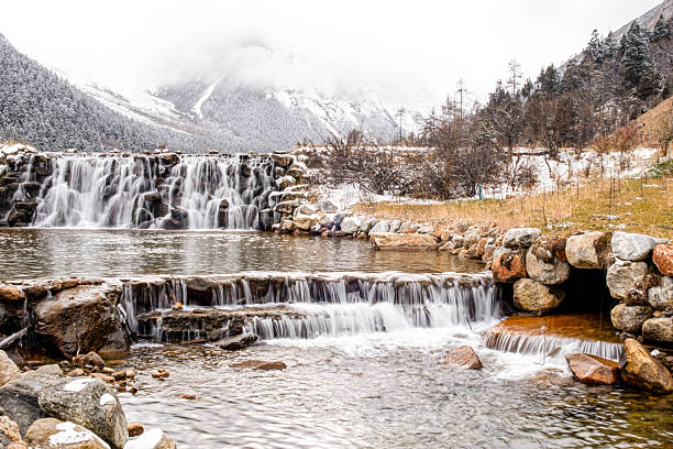 A stunning winter scenery of a cascading waterfall flowing through rocky steps, surrounded by snow-covered mountains, frosted trees, and misty clouds in a cold alpine valley.