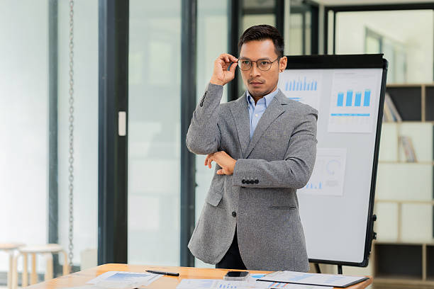 istock A confident, bespectacled young Asian businessman stands facing forward at a desk with a whiteboard behind him in his office. 2250847003