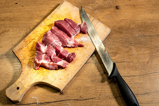 Slices of raw pork are placed on a wooden cutting board with a serrated-edge knife nearby. The cutting board has a light wood finish and visible grain patterns. The knife has a black handle, and the blade is stained with meat juices. The background is a wooden surface with a natural texture. This setup is suitable for preparing meat in a kitchen setting.