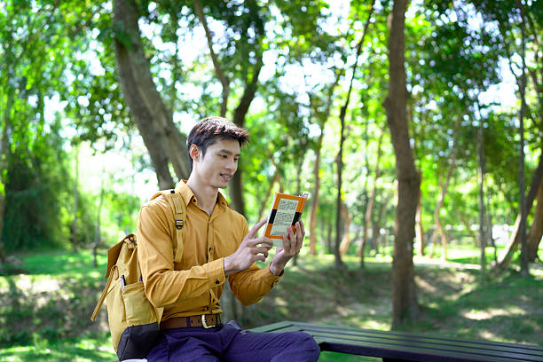 young man enjoying leisure time on a park bench, reading a book and drinking coffee on a sunny day outdoors.