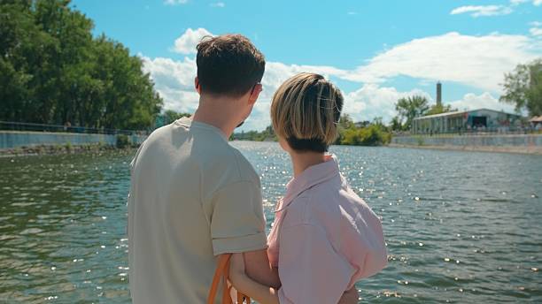White man and Asian woman walking with their white poodle along a sunny Montreal riverside dock, enjoying summer weather together.