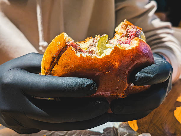 close-up shot of a partially eaten gourmet beef burger (with pickles and cheese) held by hands wearing black serving gloves. high contrast, modern food concept ideal for street food, restaurant, and casual dining advertising.