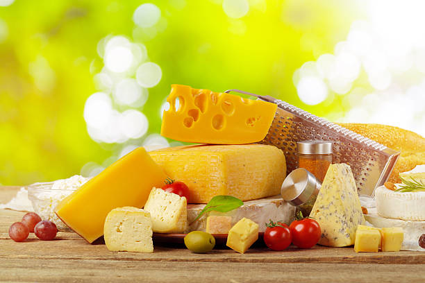 Various types of cheese arranged on a wooden table with fresh herbs and vegetables in sunlight.