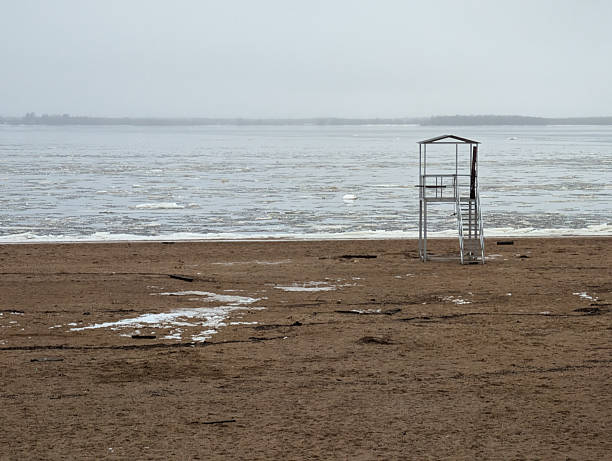 Lifeguard Chair On Icy Beach, Overcast Sky And Thin Ice Floes Drifting Near Shore Muddy Sand With Shallow
