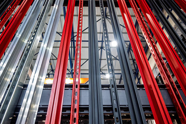 This low-angle, abstract close-up shot captures the intricate vertical elements of a modern industrial shelving system. The composition showcases a dynamic array of vibrant red, sleek silver, and dark gray metal uprights, each featuring characteristic perforations that add texture and detail. The perspective emphasizes the repetitive patterns and metallic sheens, highlighting the robust and organized nature of contemporary warehouse infrastructure. Bright ceiling lights are subtly visible in the background, reinforcing the industrial setting. This image is perfect for illustrating themes related to logistics, storage solutions, manufacturing processes, supply chain management, and modern industrial design, providing a compelling visual for corporate communications, advertising, or editorial content.