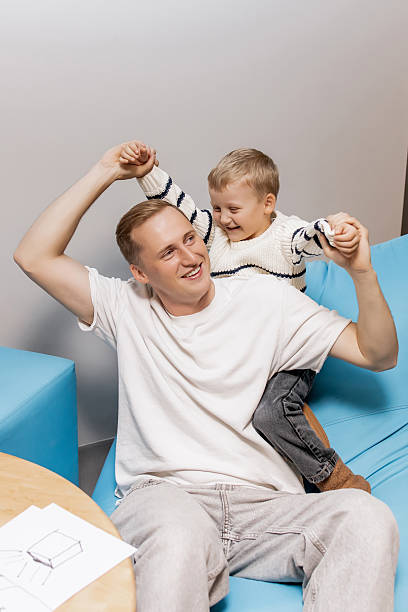 istock Joyful Father and Son Hugging and Laughing Together While Sitting on a Blue Bean Bag Chair at Home, Concept of Happy Childhood and Family Affection in Cozy Interior, Father's Day 2250914021